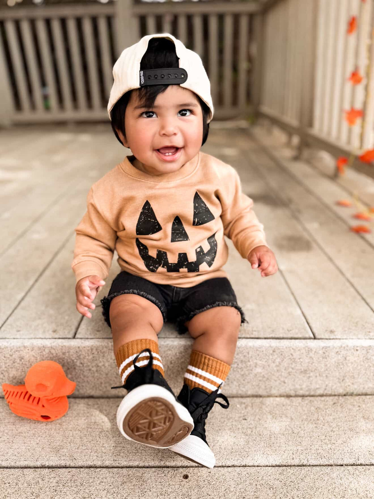 Child wearing a Halloween-themed outfit with a pumpkin face shirt on a wooden deck.