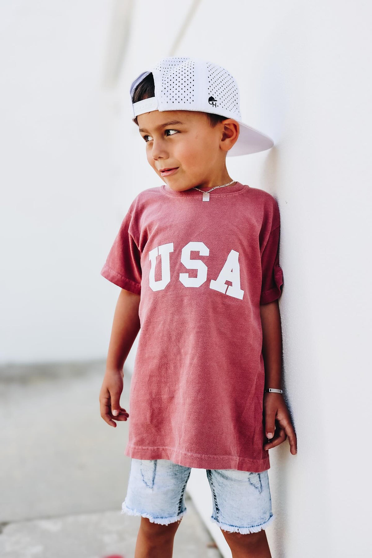 Toddler boy wearing red USA patriotic shirt with collegiate lettering, trendy boys Fourth of July outfit for kids summer celebrations.