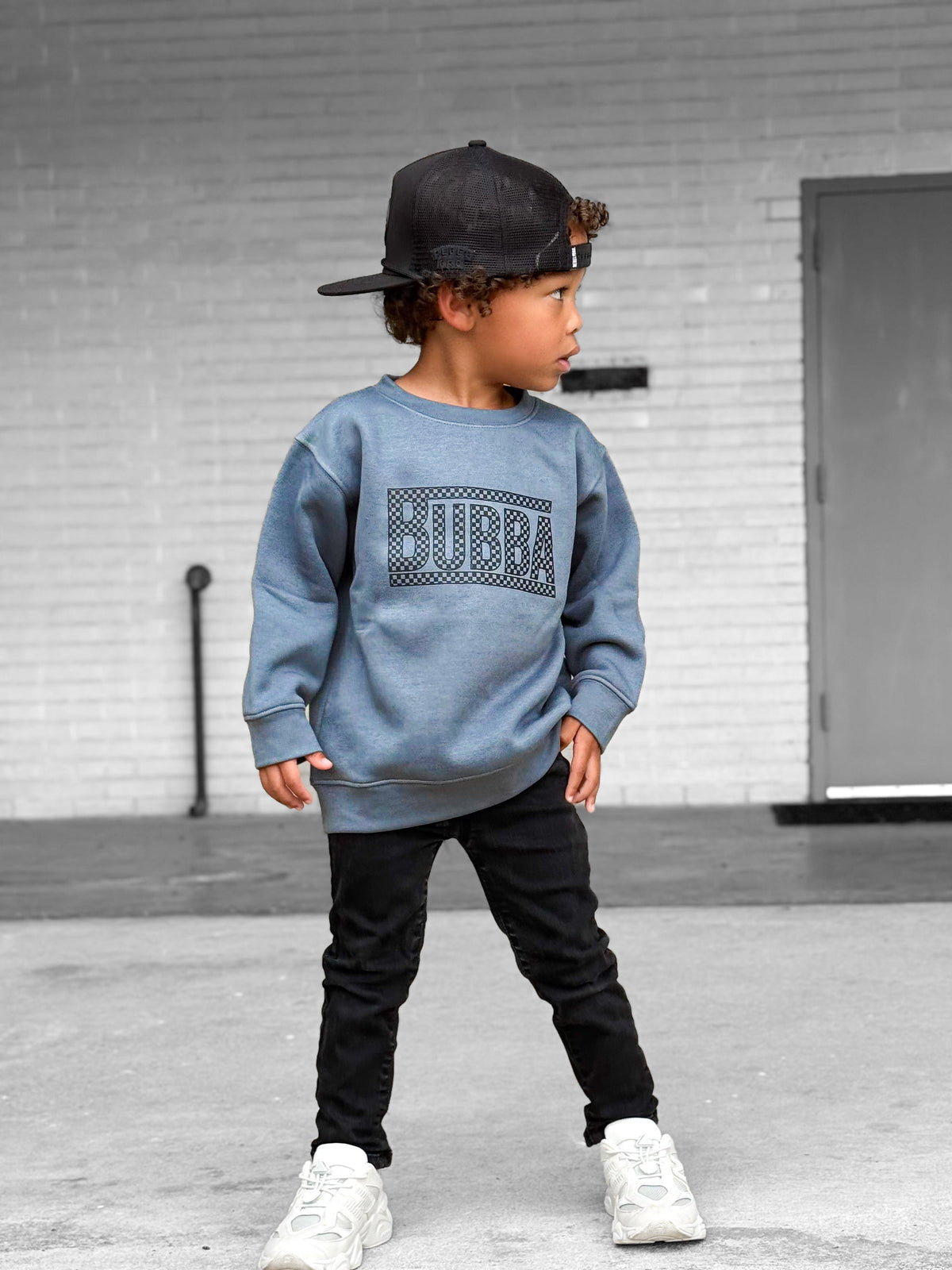 Child wearing a blue sweatshirt with 'Bubba' branding, black pants, and white sneakers standing on a concrete surface.