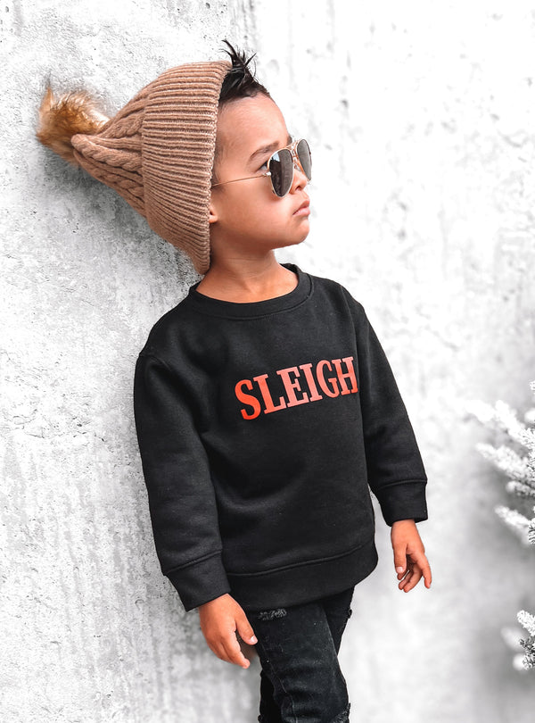 Child wearing a black sweatshirt with 'SLEIGH' text, standing next to a decorated Christmas tree.