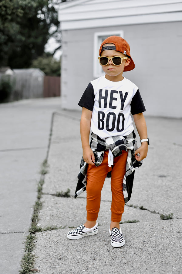 A child wearing sunglasses and a 'HEY BOO' black t-shirt with a graphic on the front, paired with orange pants and checkered shoes, standing on a sidewalk.