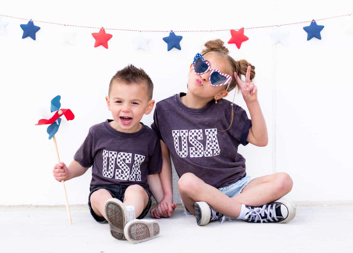 Two children sitting in front of a backdrop with red and blue stars, holding pinwheels and wearing navy-colored T-shirts with the text 'USA' on them.