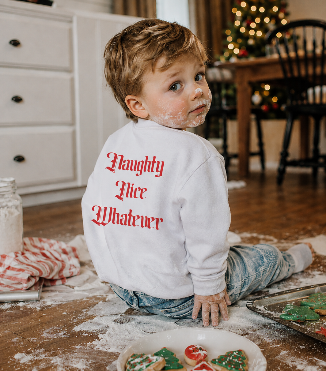 A young child sitting on a wooden floor covered in flour, wearing a white sweatshirt with 'Naughty Nice Whatever' printed in red gothic lettering on the back. The child has sugar dusted face and hands, with cookies on a tray and a plate nearby in a cozy, decorated holiday kitchen setting.