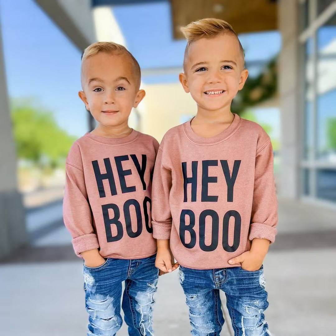 Two young boys wearing matching dusty rose long-sleeve shirts with 'HEY BOO' printed in black across the front, paired with distressed blue jeans, standing outdoors in front of a modern building.